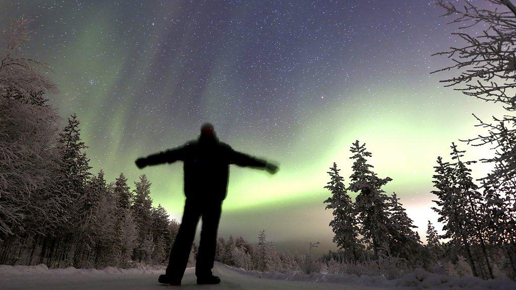 Pessoa vestida com roupas de frio em meio a uma paisagem coberta de neve, com uma aurora boreal verde visível no céu noturno