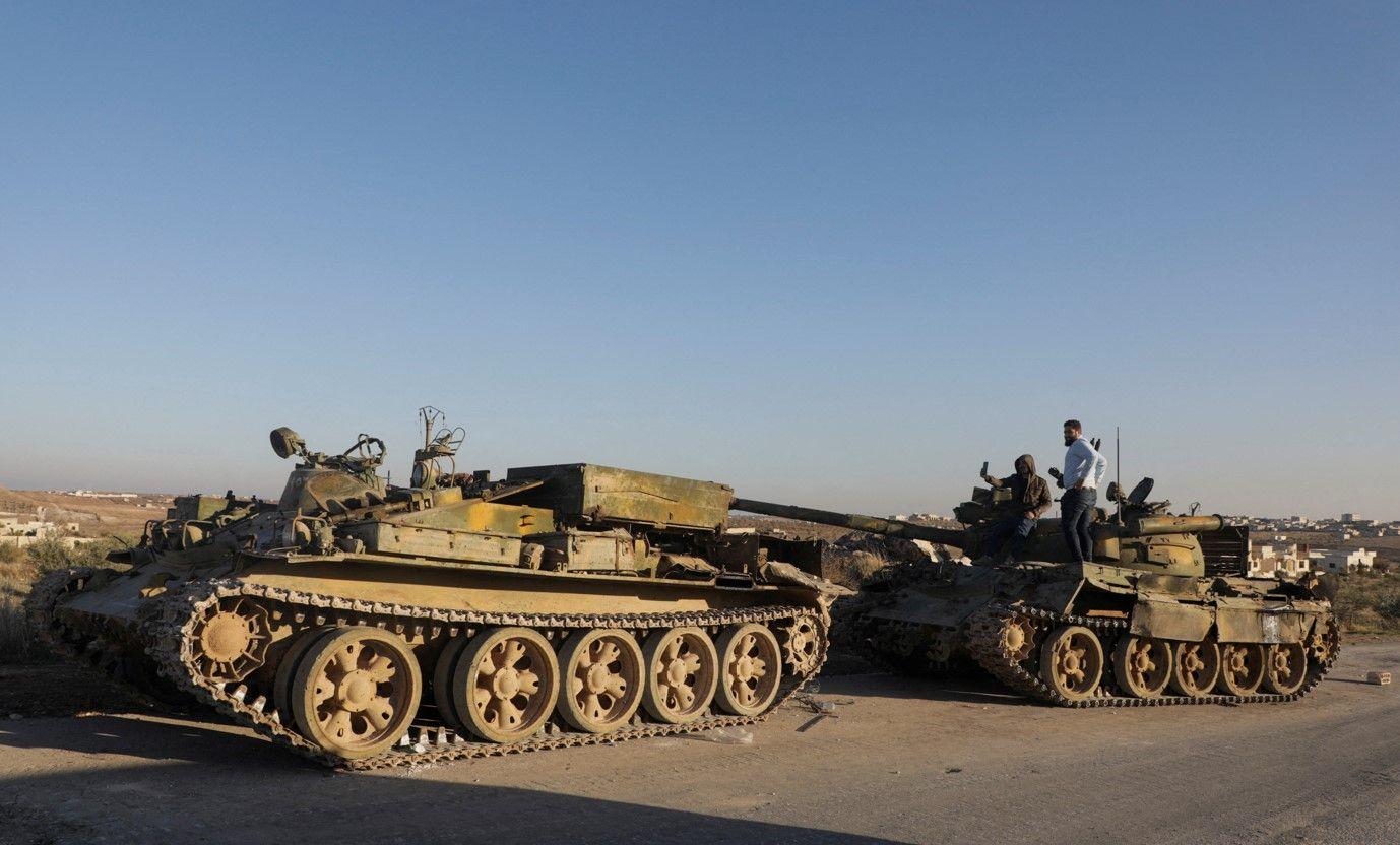 أشخاص يقفون على مركبة عسكرية متضررة في مدينة معرة النعمان بمحافظة إدلب، بعد استيلاء المعارضة على المدينة.