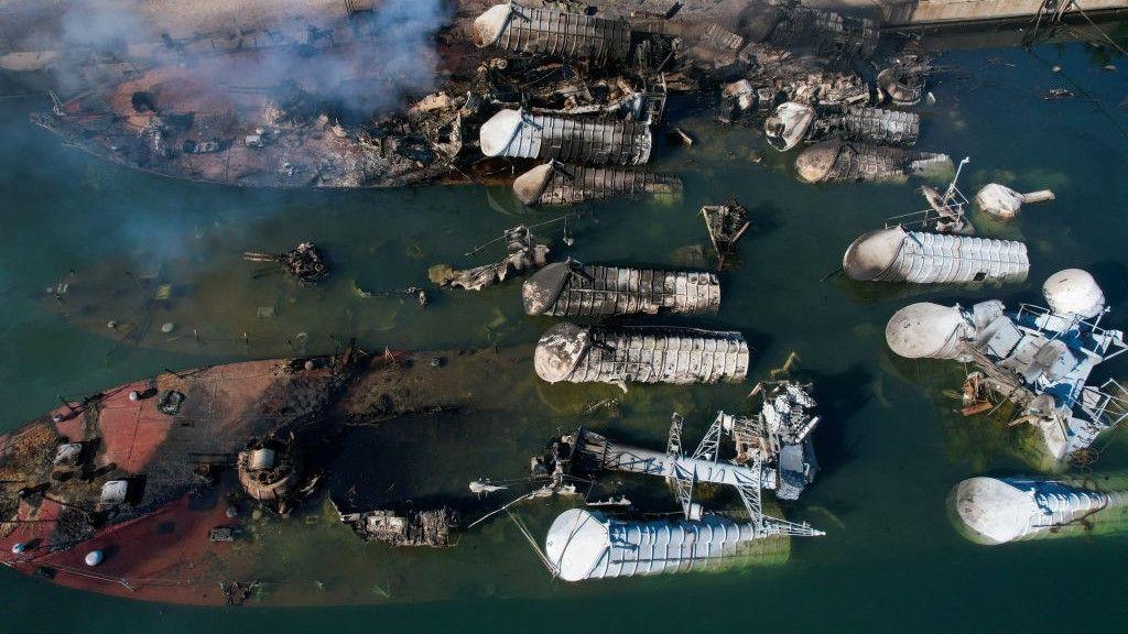 Uma foto aérea de vários navios afundados perto do cais com fumaça subindo.