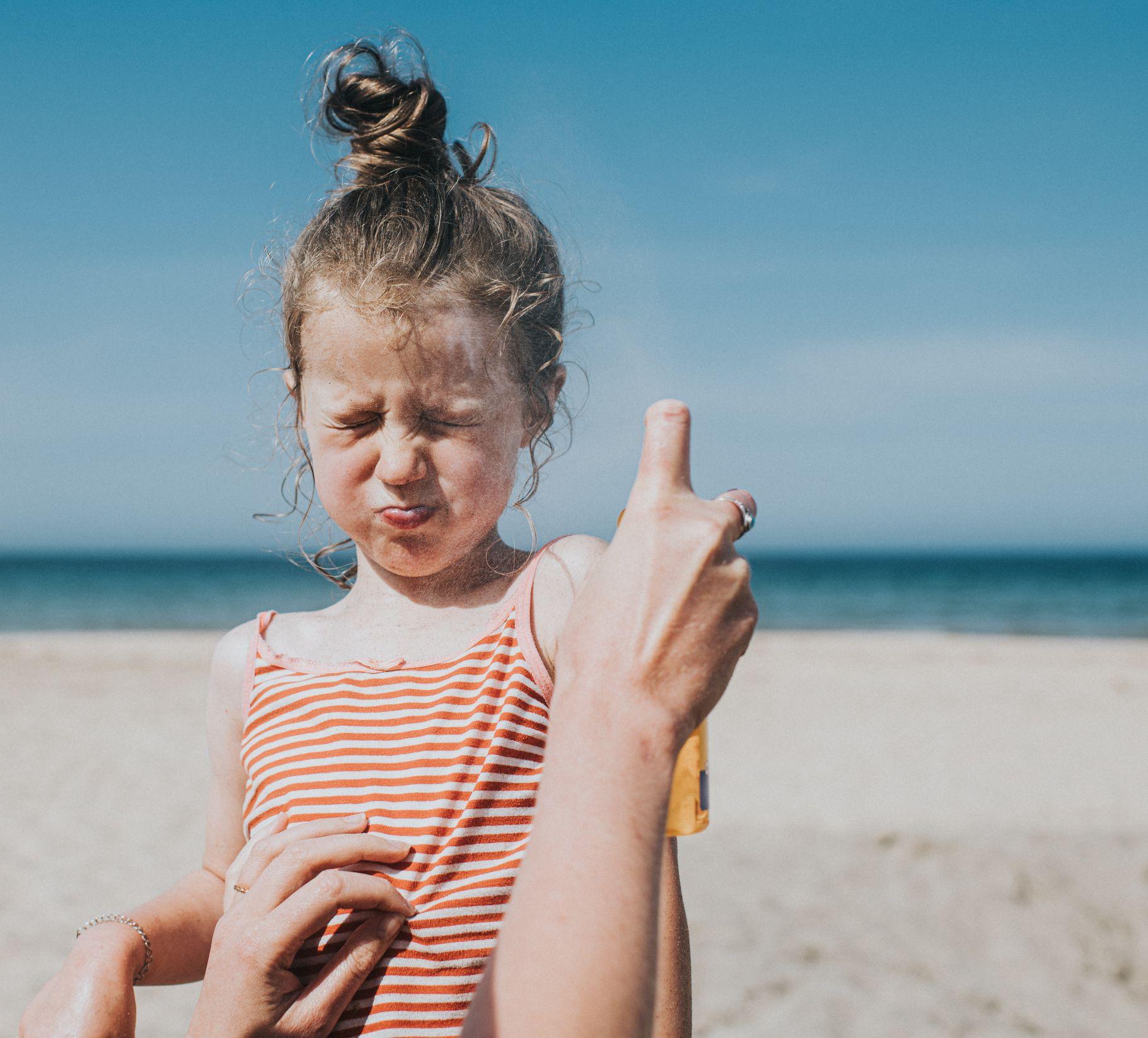 فتاة صغيرة لطيفة تحبس أنفاسها وتجفل بينما تقوم سيدة تظهر يداها في الصورة، برش واقي الشمس على وجه الطفلة الصغير، أثناء وقوفهما على الشاطئ.