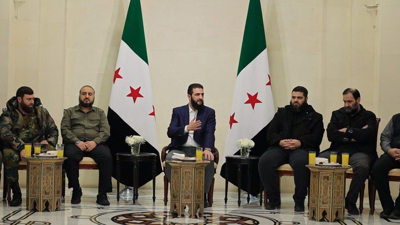 صورة تظهر قائد هيئة تحرير الشام أحمد الشرع داخل قاعة وحوله عدد من ممثلي الفصائل المسلحة في سوريا.