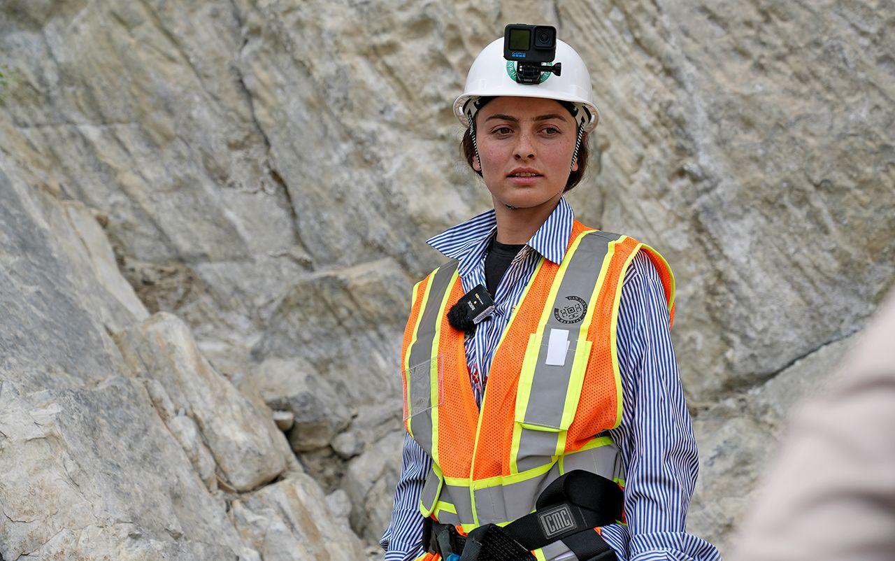 A head and shoulders shot of an Aga Khan Agency for Habitat worker wearing high vis over a stripy shirt and a hard hat helmet with a GoPro on top with a rockface in the background