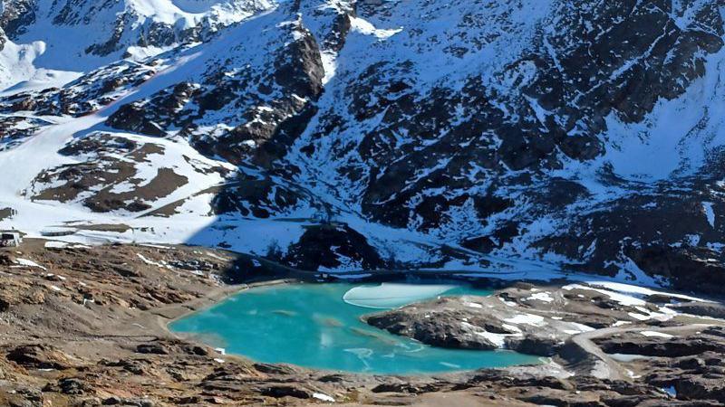 A imagem mostra a geleira Hochjochferner no outono, parcialmente derretida, com um lago azul-turquesa no centro, cercado por montanhas cobertas de neve e terreno rochoso exposto.