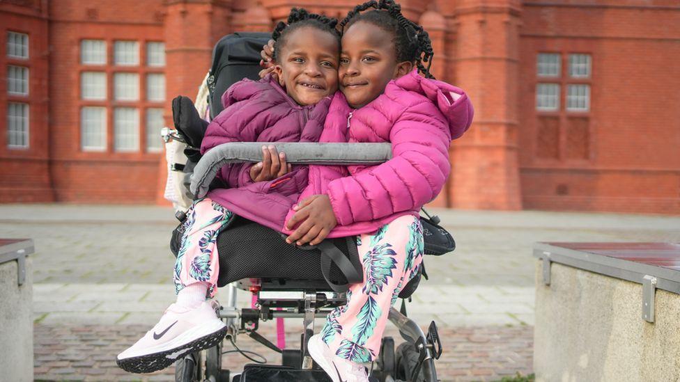Gêmeas siamesas de 7 anos, negras e com trancinhas nos cabelos, sentadas em um carrinho, com roupas de frio, sorrindo