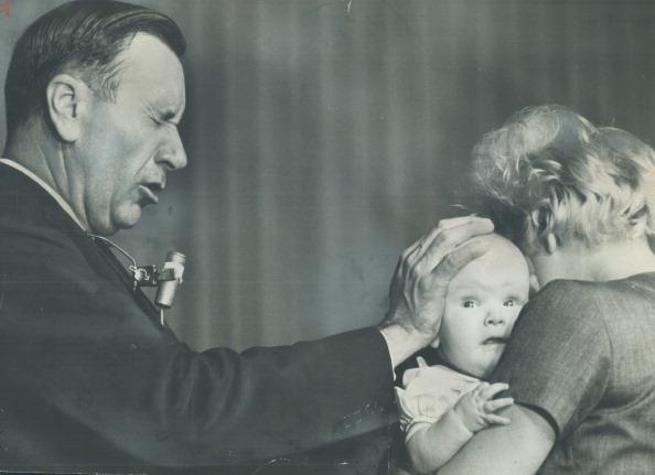 O evangelista Oral Roberts com a mãe sobre a cabeça de um bebê, que está no colo da mãe