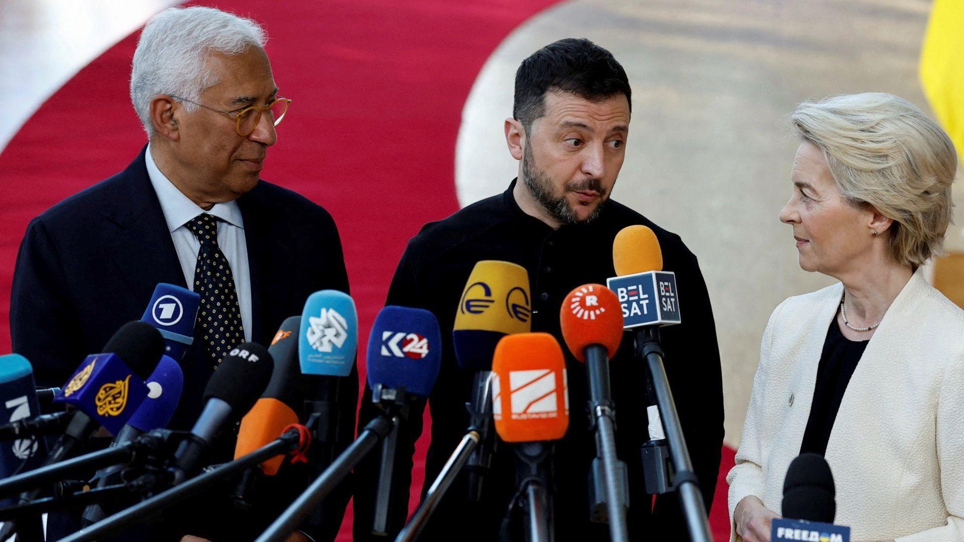 Volodymr Zelensky dressed in black fatigues stands at a podium with several mics in front of him. He looks at Ursula Von Der Leyen as Antonio Costa stands behind him. 