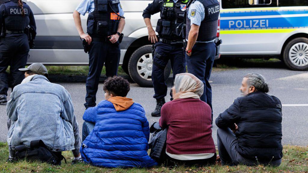 Varias personas esperan sentadas en una cuneta mientras policías alemanes hablan de pie.