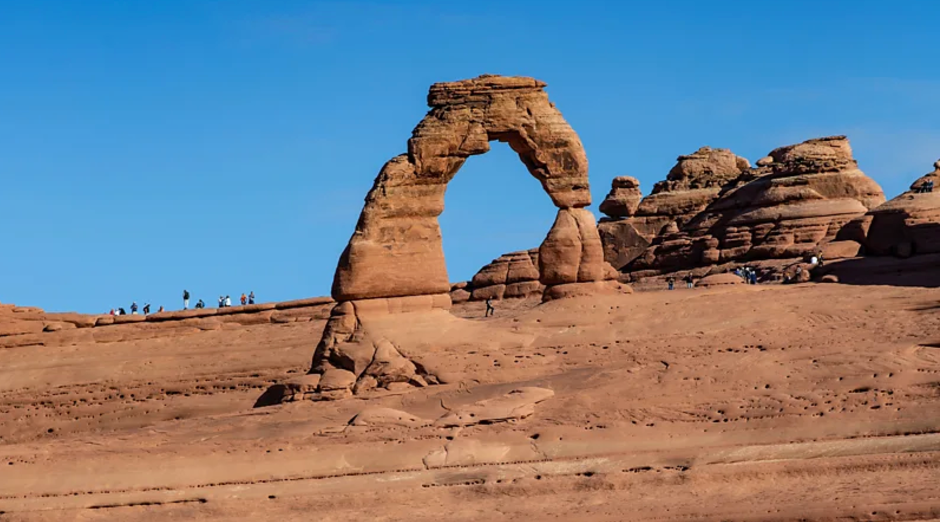 Um arco natural de rocha arenosa em um deserto, com pessoas em pé ao redor e um céu azul ao fundo