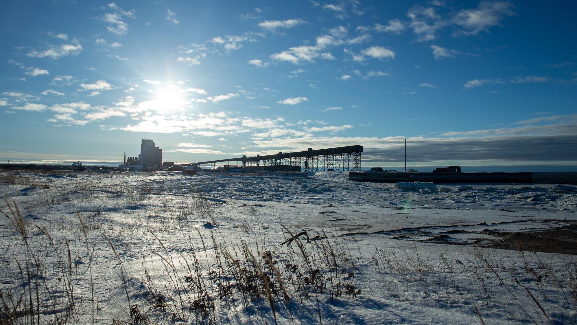 A imagem mostra Churchill Harbour em um ambiente coberto de neve e gelo, com céu aberto e sol baixo no horizonte. A estrutura do porto se destaca no cenário, que fica fechado nove meses do ano por causa do gelo.