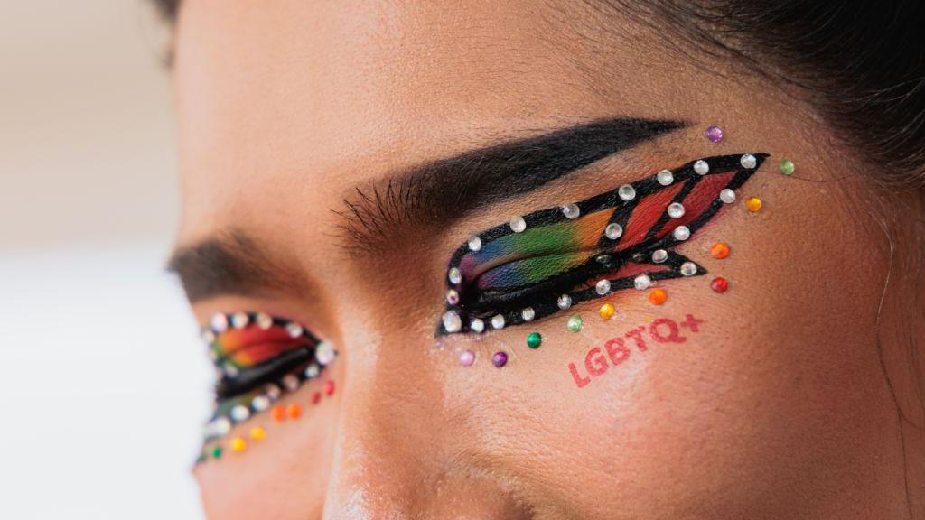 Mulher com sombra e maquiagem nas cores do arco-íris, ostenta as letras LGBTQ+ em vermelho abaixo dos olhos fechados