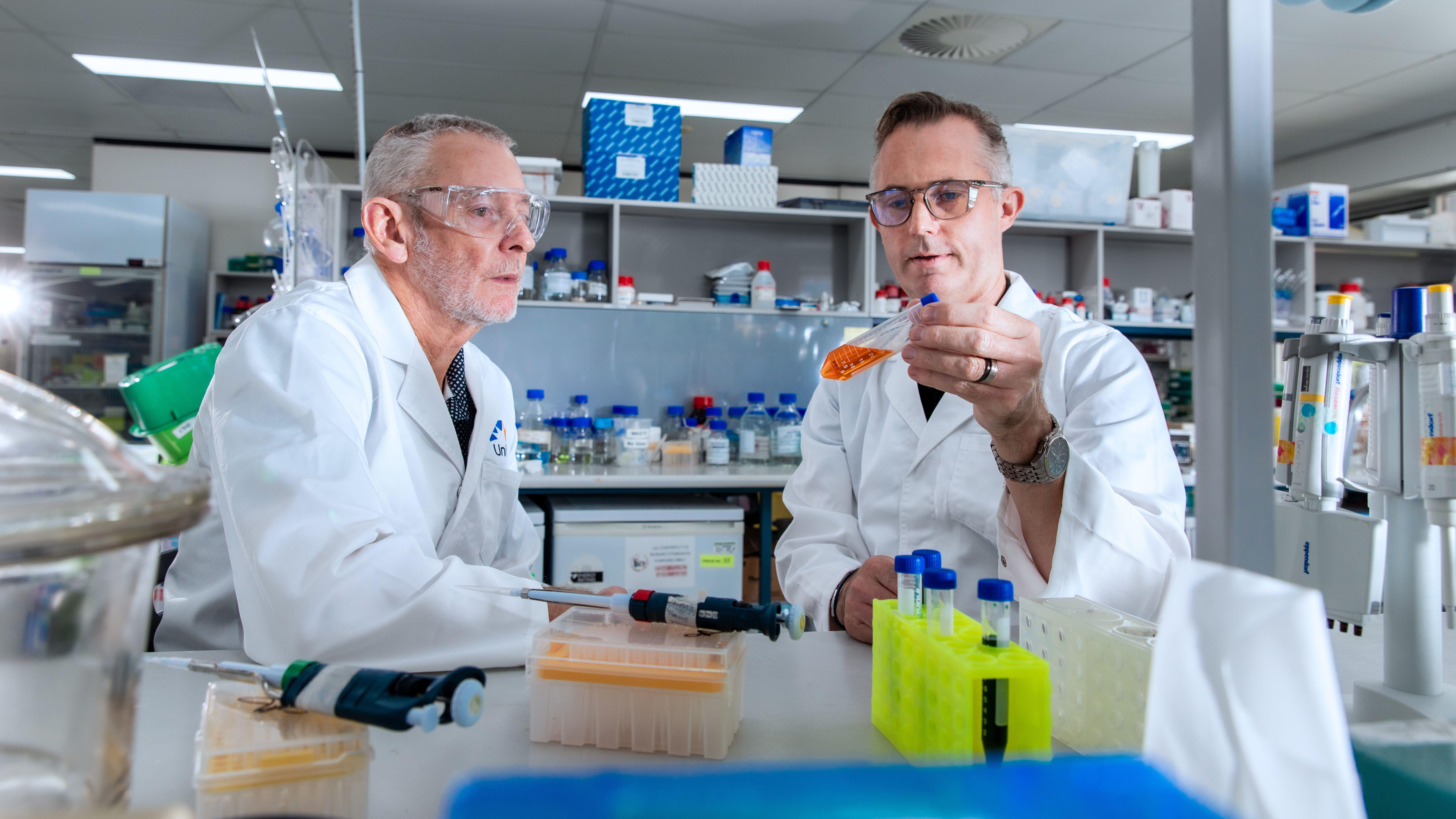 Two men examine a test tube with orang liquid inside while sitting in laboratory. They are wearing safety goggles and white coats.