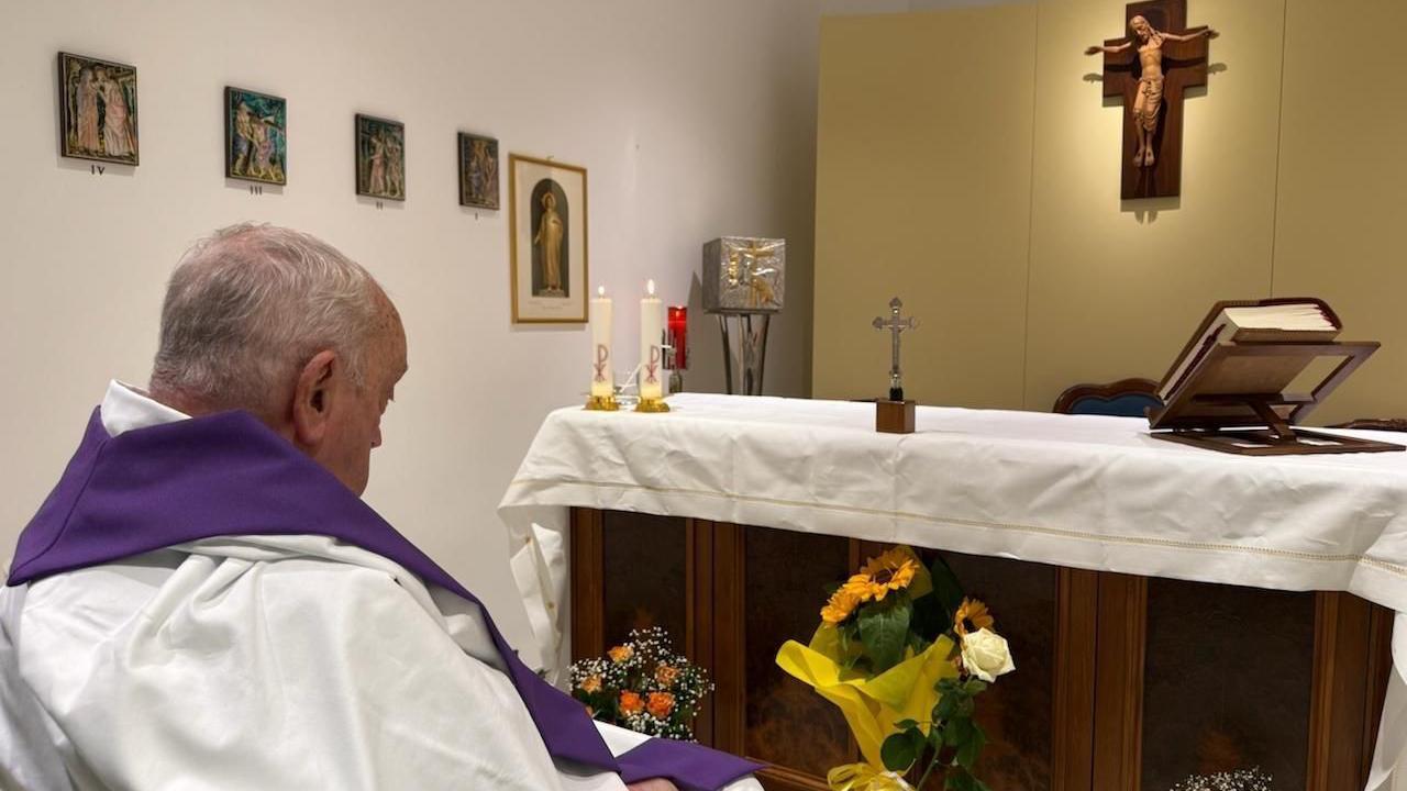 El Papa, sentado en silla de ruedas, observa un altar en una habitación pintada de blanco. Hay una Biblia, un crucifijo y algunas velas en el altar, y una representación de Cristo en la cruz cuelga sobre él.