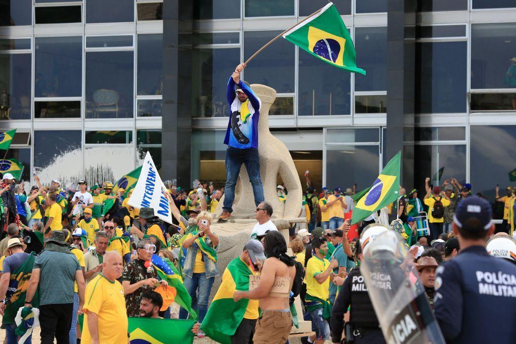 Centenas de pessoas ao redor da estátua da Justiça em frente ao STF; há um manifestante em pé em cima da estátua, hasteando a bandeira do Brasil