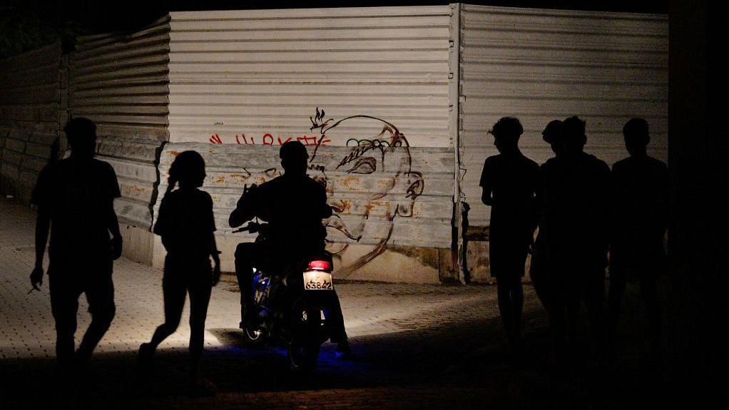 Unas personas caminan por una calle oscura en Cuba, iluminadas solamente por el faro de una motocicleta.