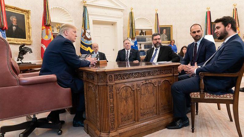 Donald Trump observa brasileiros durante reunião com equipe do presidente Bolsonaro. Seis pessoas estão sentada, usando terno, em torno de uma mesa de madeira na Casa Branca