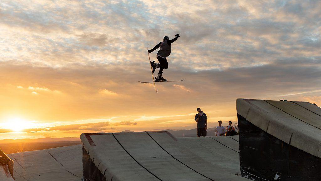 Homem manobra no ar em uma pista de esqui seca com o pôr do sol ao fundo 