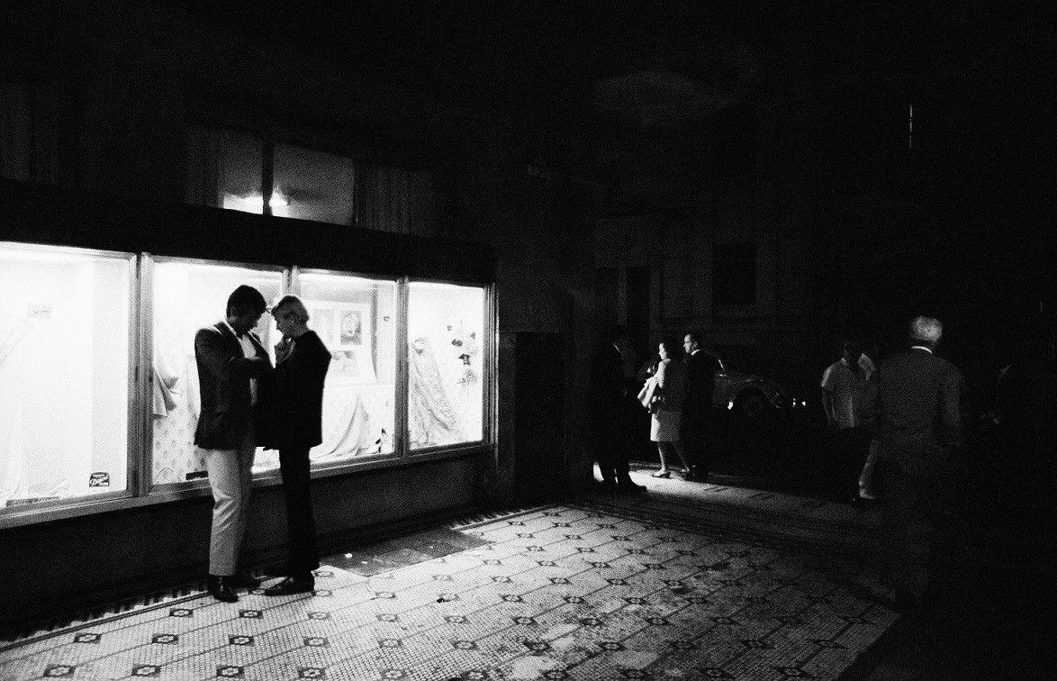 Dois homens em frente a vitrine, durante a noite; no canto das fotos, aparecem outras pessoas caminhando