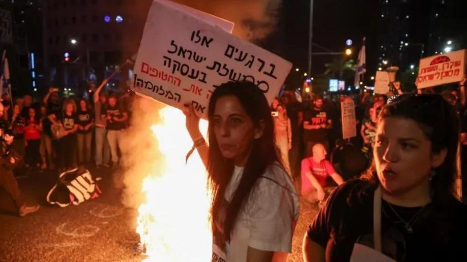 Pessoas reunidas em volta de um fogo realizam um protesto com cartazes em hebraico 