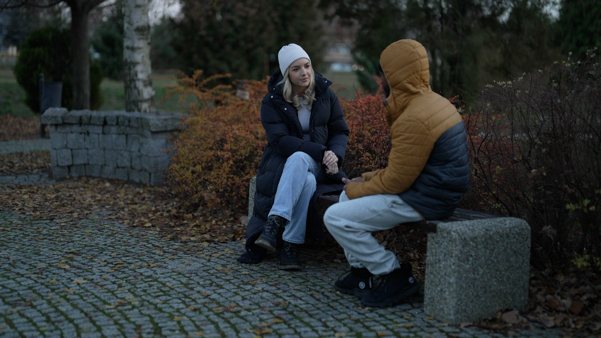 A man in grey trackpants and a hooded yellow and black jacket sits on a park bench next to a woman in a white hat, black puffer coat, jeans and boots.