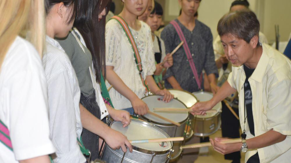 Homem de meia idade ensina jovens a tocar um instrumento de percussão. 