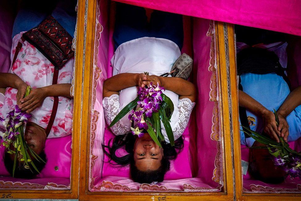 Mujeres con flores acostadas en unas cajas que parecen ataúdes