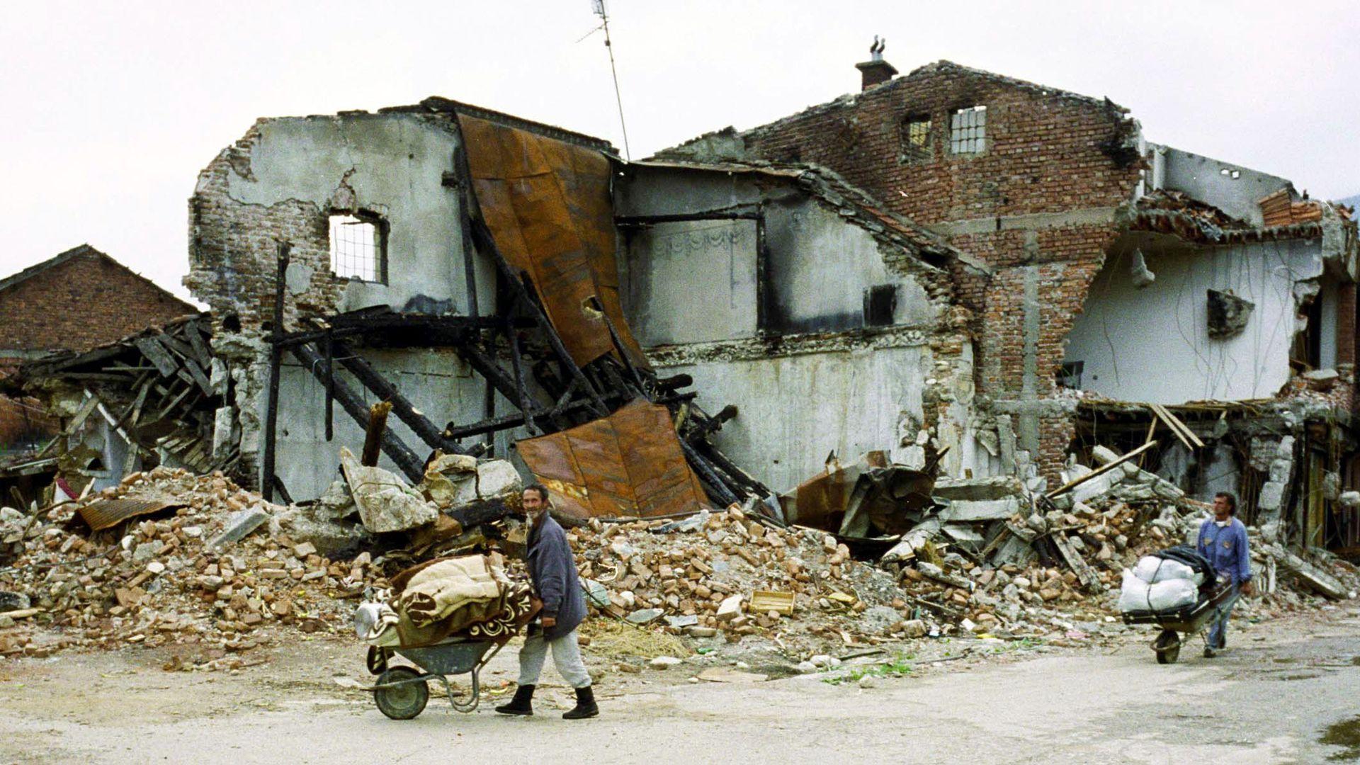 من ذوي الألبان يدفعون عربات اليد التي تحتوي على ممتلكاتهم أمام مبنى مدمر، أبريل 1999