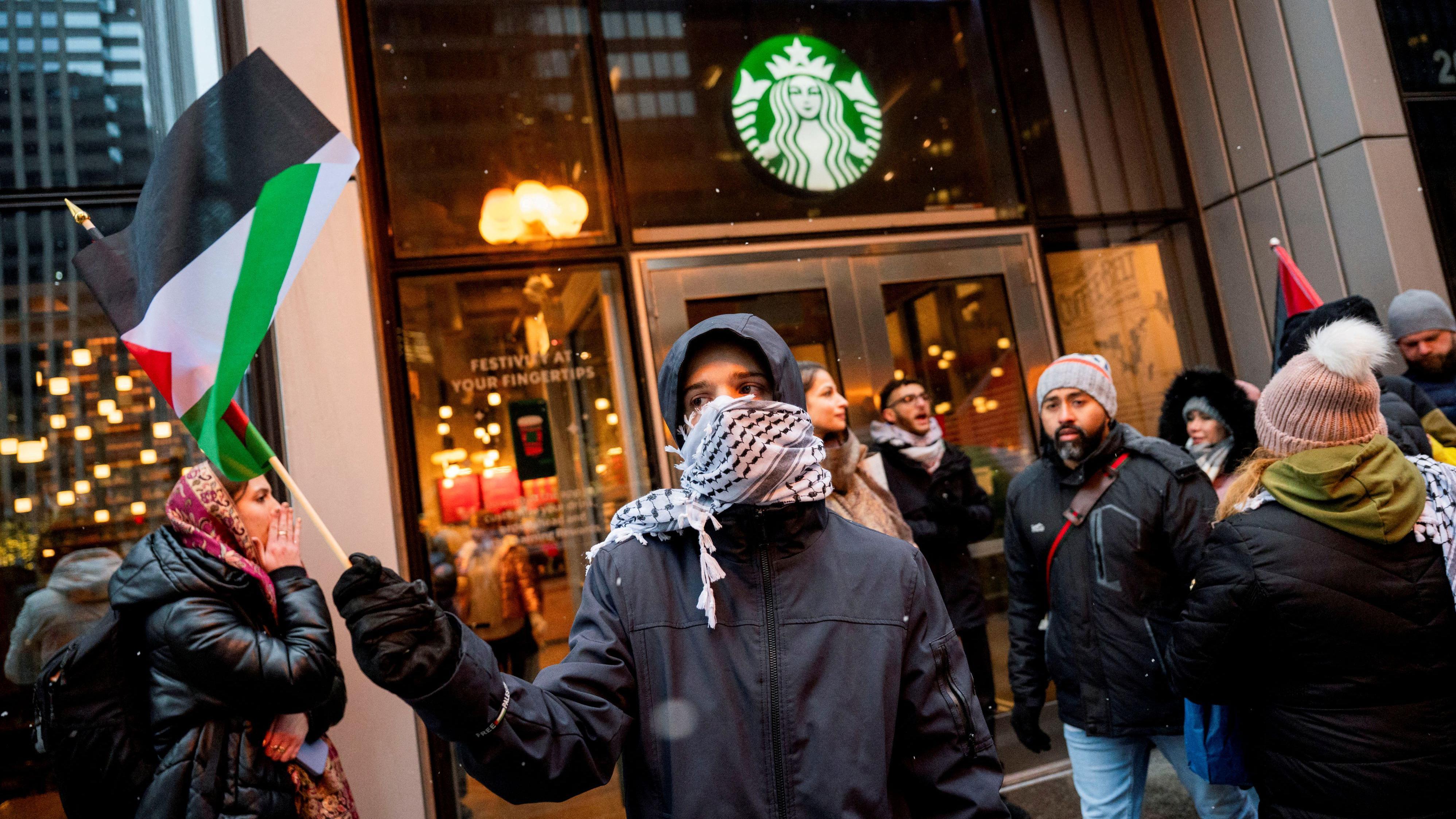 A imagem mostra um protesto em frente a uma loja da Starbucks. No primeiro plano, um homem de casaco preto, com um lenço branco e preto no rosto, segura uma bandeira pequena da Palestina.