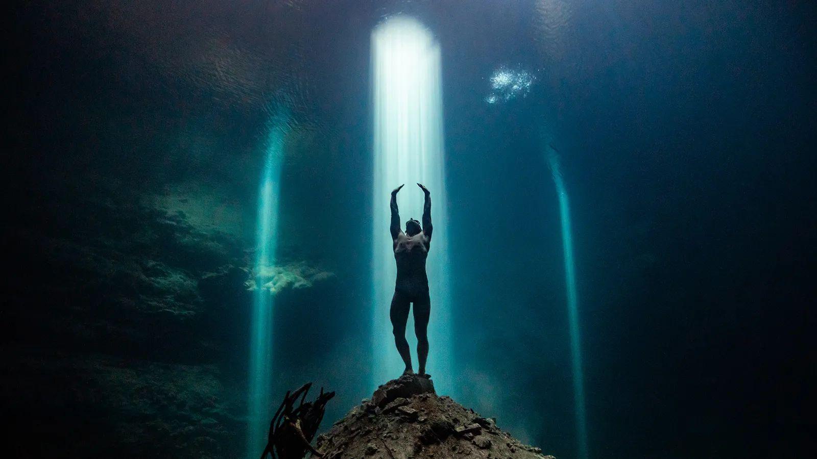 Hombre bajo el agua parado en un montículo de piedra bajo un rayo de luz en cenotes de méxico