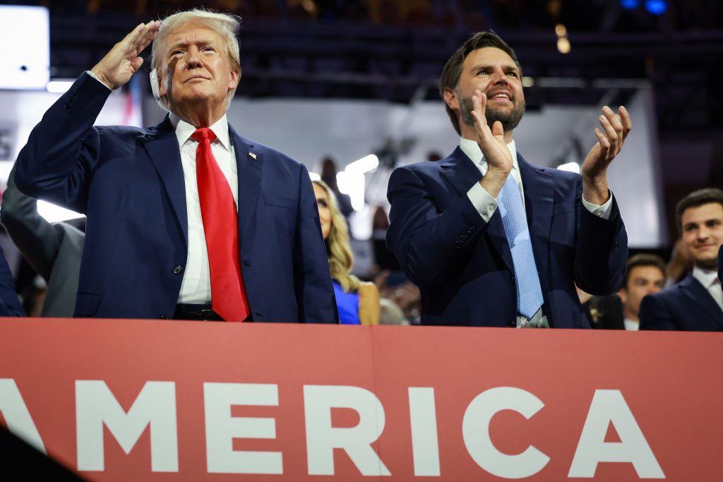 Trump e JD Vance em pé, sorrindo, durante um grande evento, cercados por outras pessoas