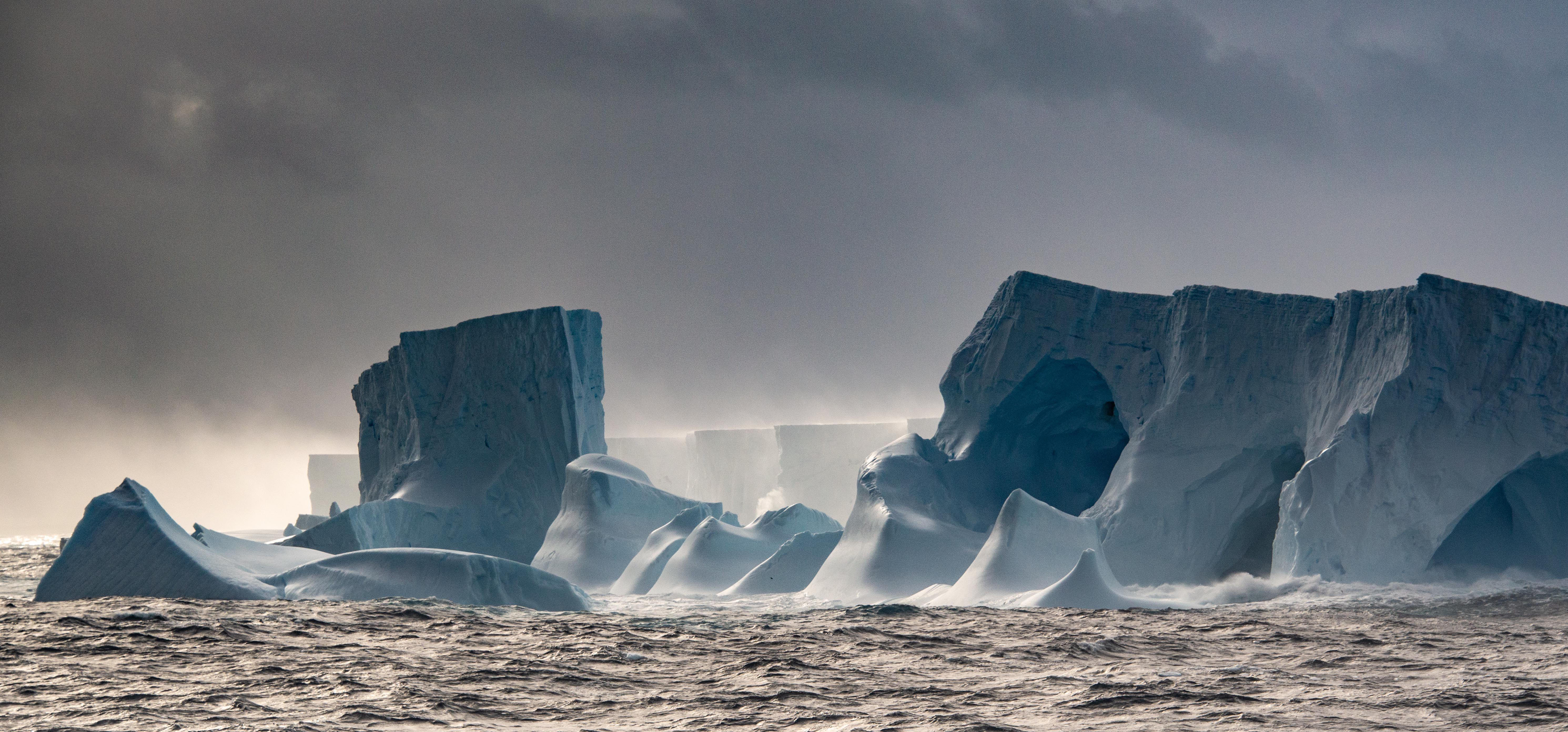 A foto mostra o imenso iceberg no mar, sob um céu cinza 