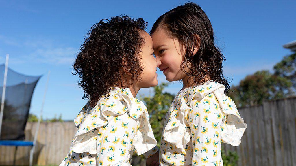 Fotografia colorida mostra duas meninas vestindo as mesmas roupas tocando seus rostos uma na outra