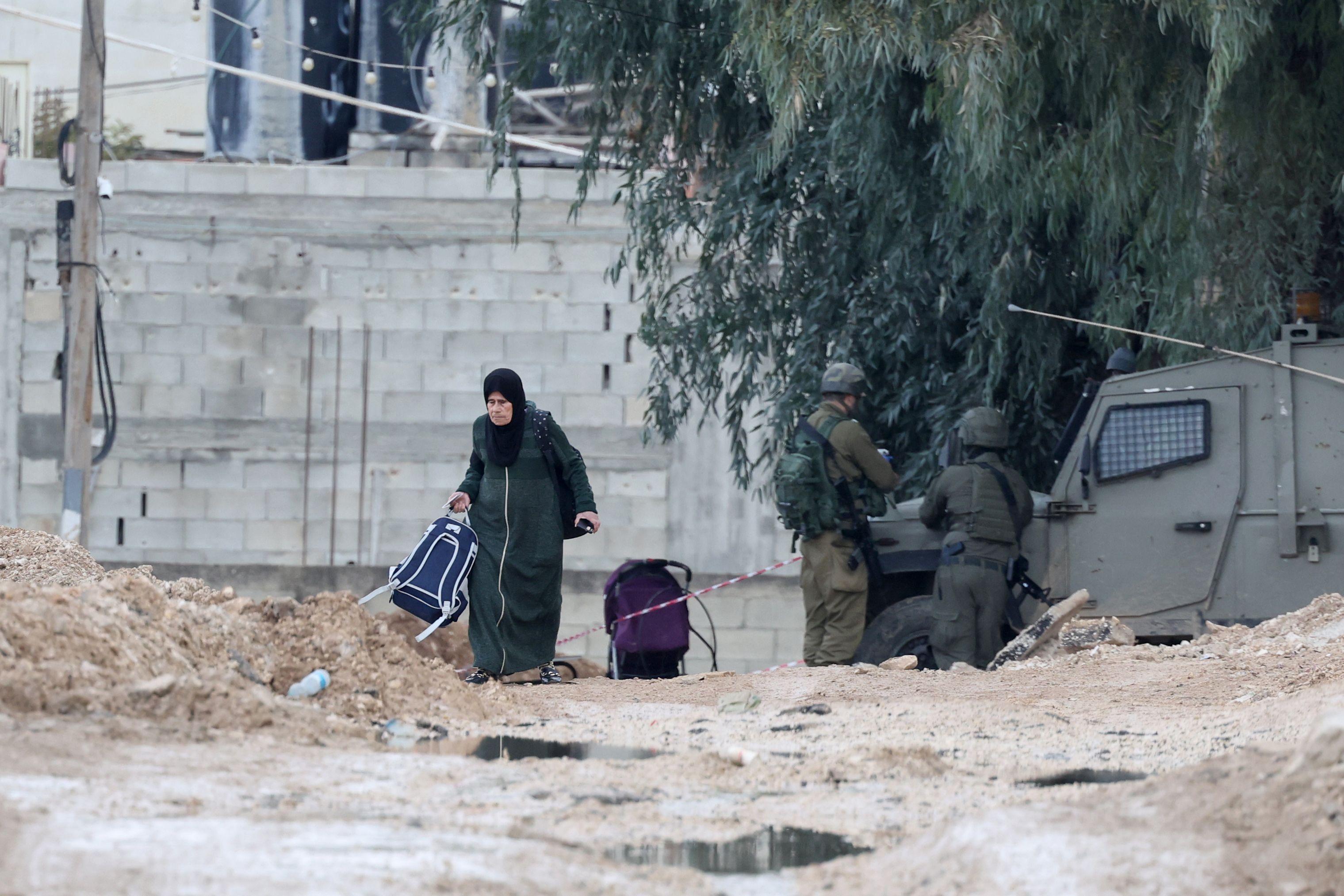 سيدة فلسطينية تحمل أمتعتها تمر أمام جنود إسرائيليين أثناء مغادرتها مخيم جنين للاجئين في اليوم الثالث من العملية العسكرية الإسرائيلية في مدينة جنين بالضفة الغربية، 23 يناير 2025.