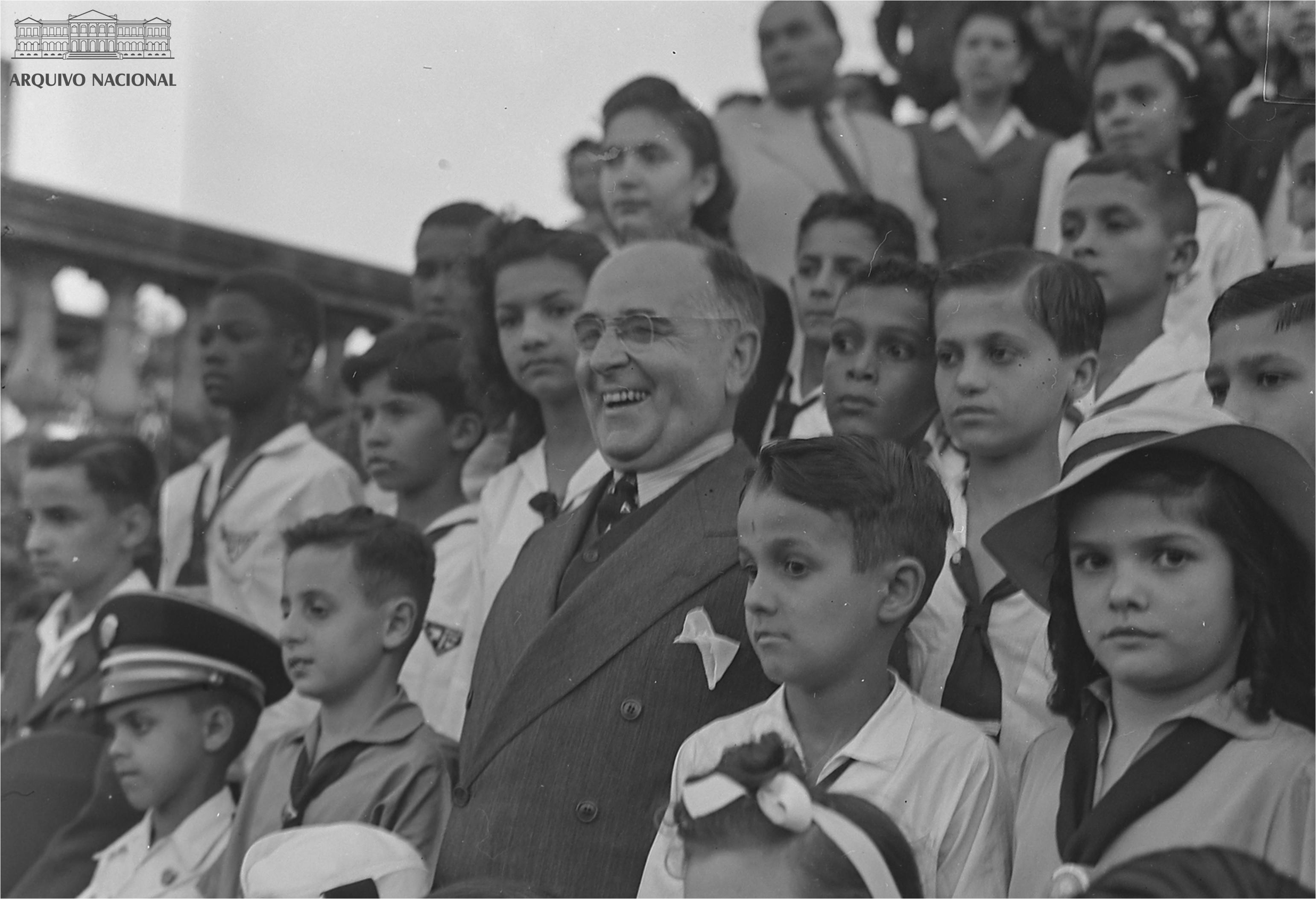 Foto em preto e branco de Getúlio Vargas cercado de crianças. Ele usa terno cinza, está de óculos e sorri. As crianças vestem o uniforme escolar da época e estão sérias 