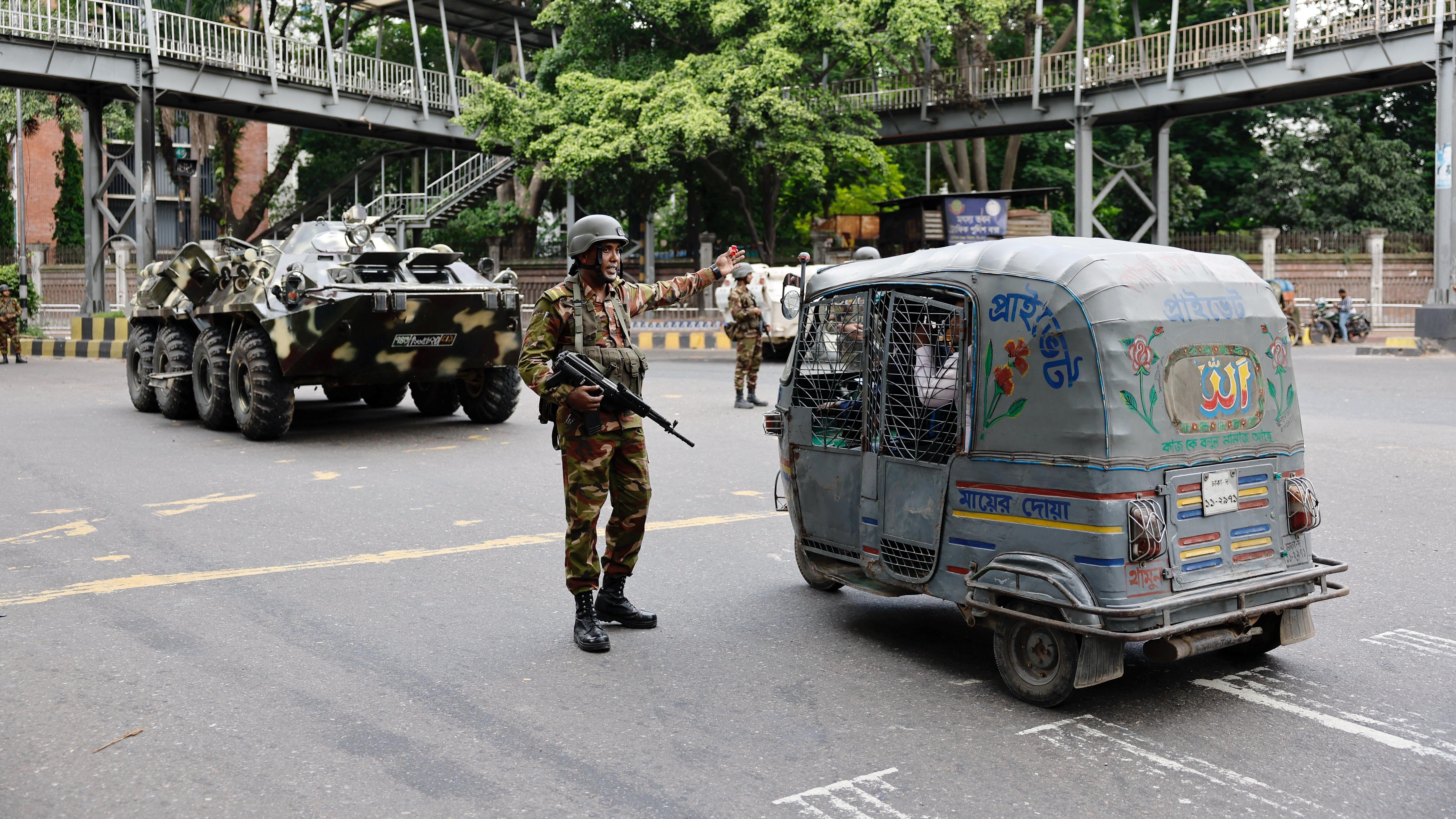 Um homem com roupas militares está na estrada perto de um tuktuk com um tanque atrás dele.