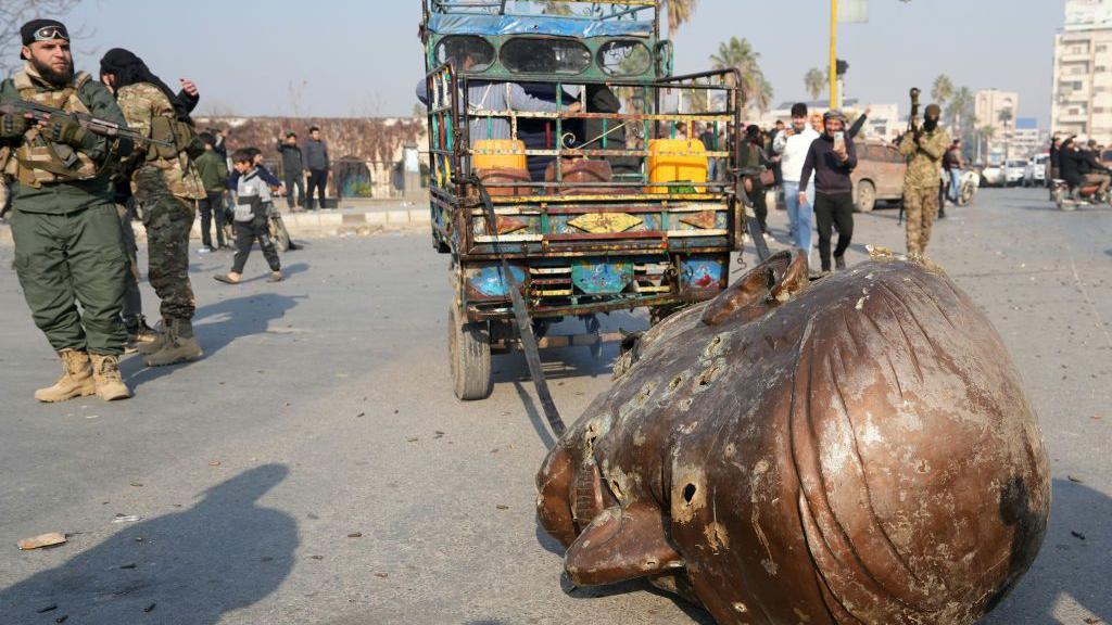 Uma caminhonete arrasta a cabeça derrubada de uma estátua do falecido presidente sírio Hafez al-Assad pelas ruas da cidade de Hama, no centro-oeste do país, em 6 de dezembro de 2024.