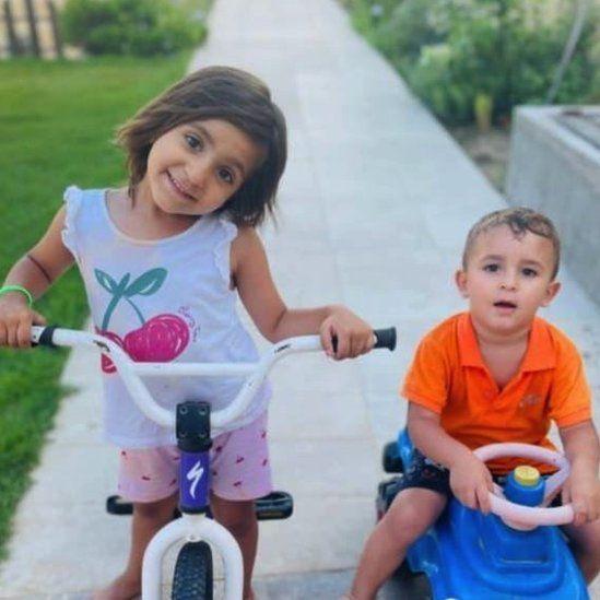 Una niña en bicicleta y un niño en un camión de juguete juegan en un jardín.