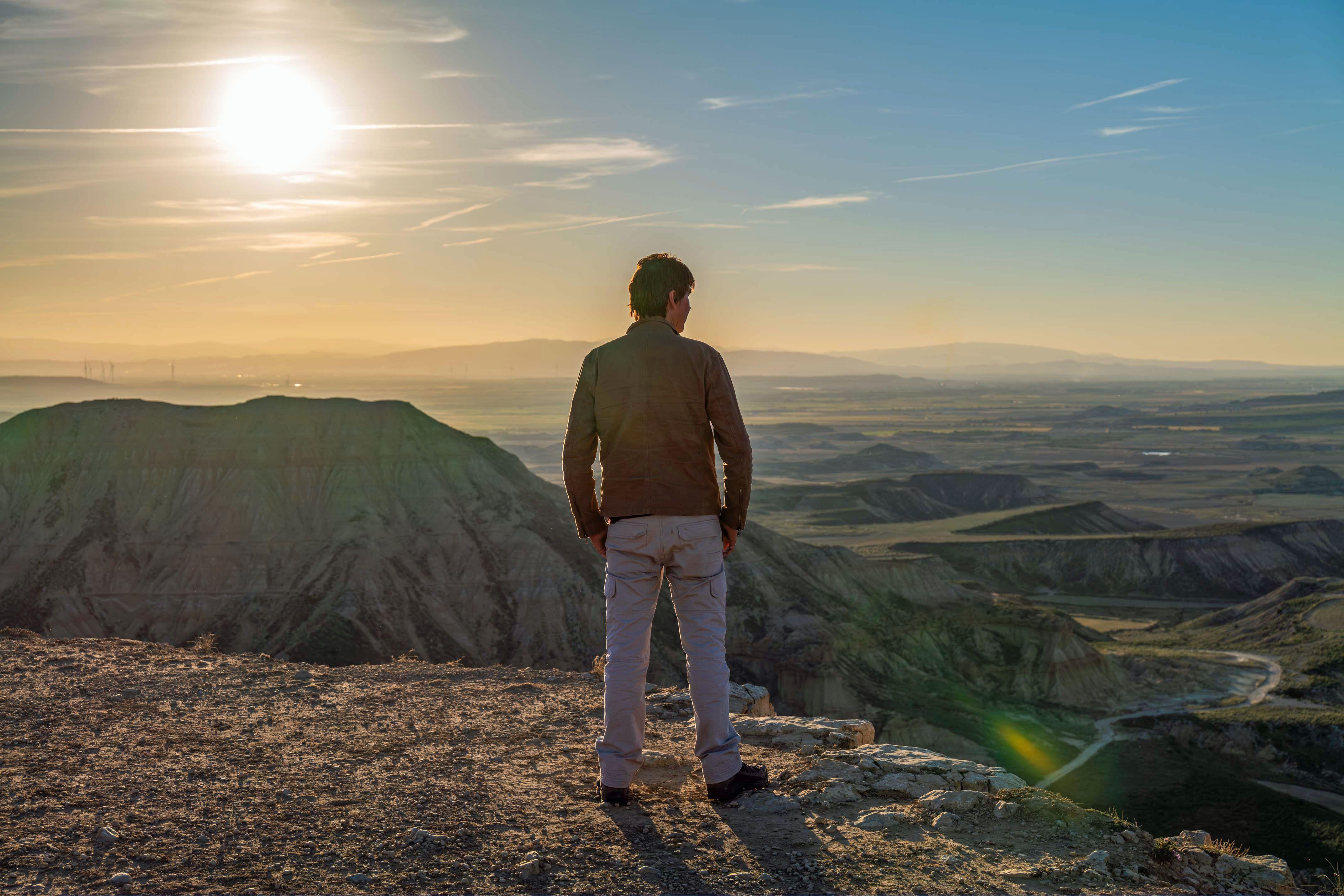 O Prof. Brian Cox olha o horizonte do topo de uma montanha. Ele está vestindo uma jaqueta marrom e calça cinza. Colinas verdes podem ser vistas à distância e o sol brilha intensamente