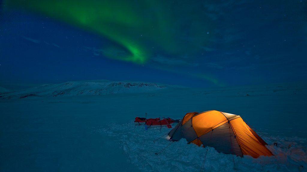 Uma barraca de acampamento em meio à neve com uma aurora boreal verde visível no céu noturno