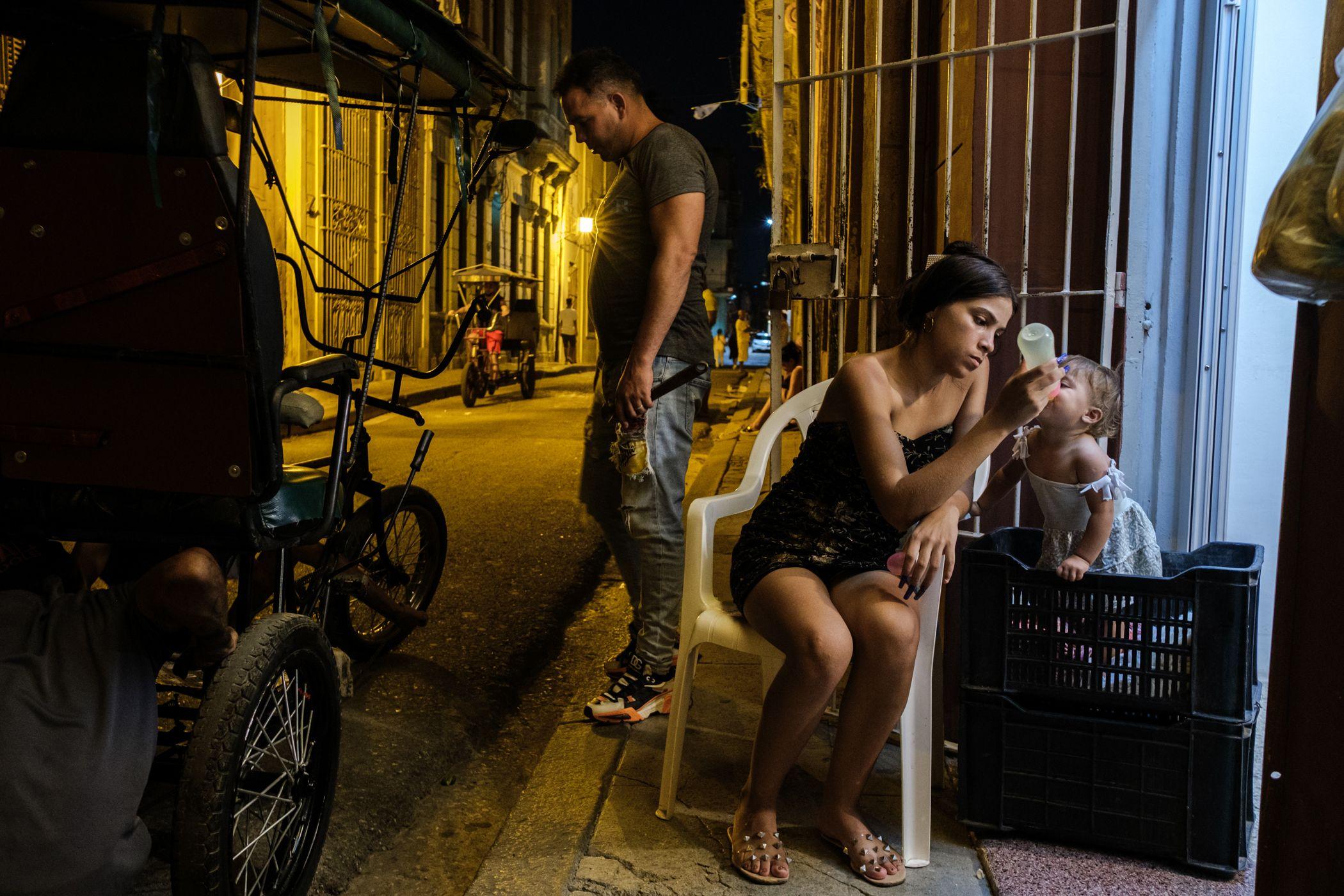 Una madre joven alimenta a su hija con un biberón, en el exterior del abasto en donde trabaja, en La Habana, Cuba. 