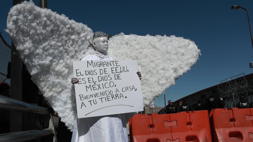 Un niño disfrazado de ángel con un cartel que da la bienvenida a los deportados.