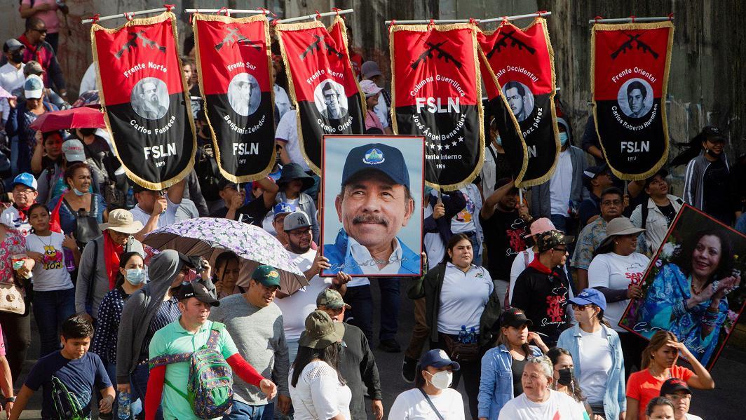 Várias pessoas na rua, algumas segurando estandartes com sigla da FSLN e uma foto de Ortega