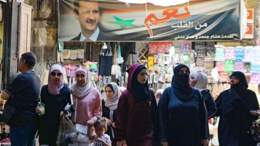 Mujeres en un mercado y arriba un cartel con el rostro del presidente sirio
