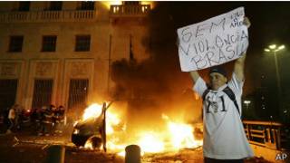 Manifestante ergue cartaz diante de veículo incendiado em frente à Prefeitura de São Paulo