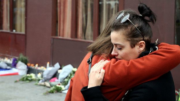 Dos mujeres se abrazan. En el fondo, el bar Le Carrillon y flores en honor a las víctimas del atentado.