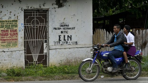 Una pareja pasa en moto por una casa con una pintada del ELN