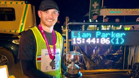 2019 Parish Walk winner Liam Parker holding the trophy with his finishing time on a screen behind