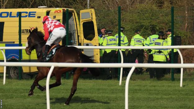 A horse running the Grand National passes by a section of fencing, behind which police are leading away protesters