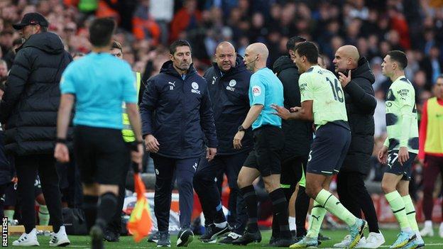 Anthony Taylor makes his way through a crowd of Manchester City players and coaches to inspect the pitchside monitor.