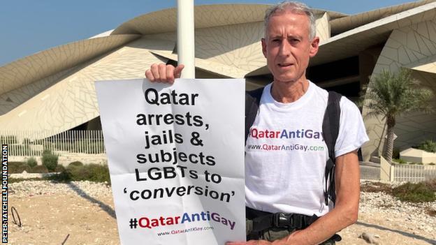 Peter Tatchell holds placard reading Qatar arrests, jails subjects LGBTs to conversion, with the hashtag #QatarAntiGay.