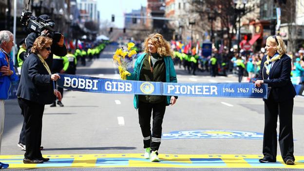Bobbi Gibb crosses a ceremonial finishline with her name emblazoned on the ribbon in 2016